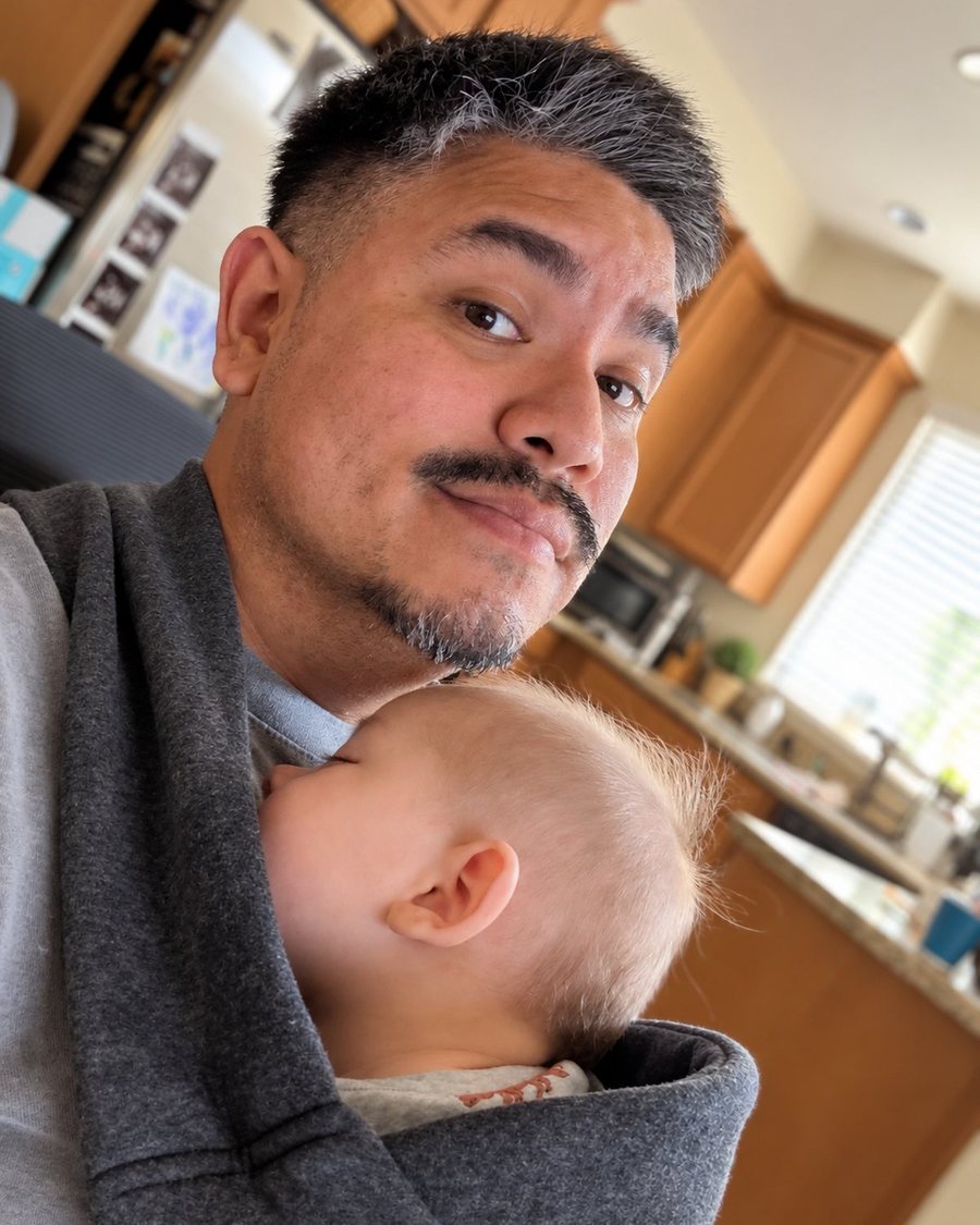 Chris at home with his youngest son asleep in a carrier
